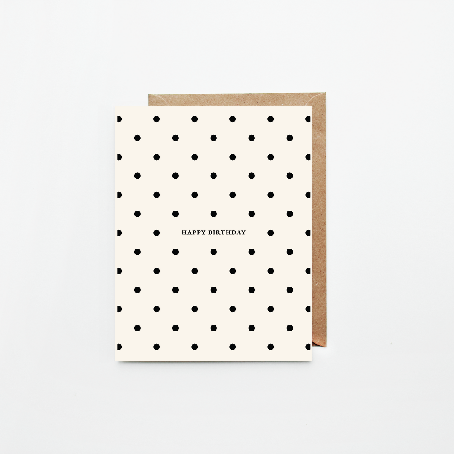 A birthday card with a black polka dot pattern on an off-white background, featuring the text 'HAPPY BIRTHDAY' in the center. It is shown with a brown kraft envelope behind it on a clean white background.