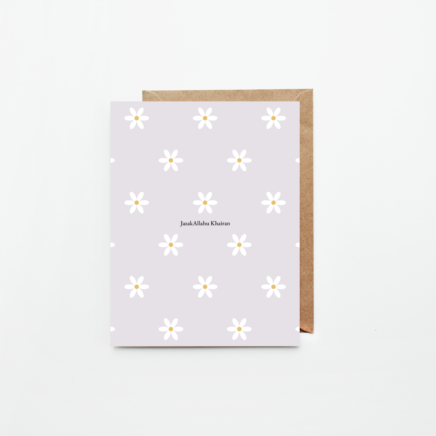 A greeting card with a light lavender background, white daisy pattern, and the name 'JazakAllahu Khairan' printed in the center, shown with a brown kraft envelope on a clean white background.