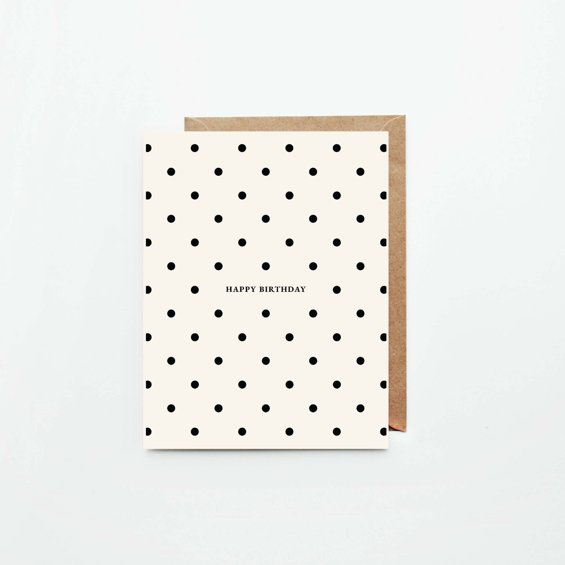 A  birthday card with a black polka dot pattern on an off-white background, featuring the text 'HAPPY BIRTHDAY' in the center. It is shown with a brown kraft envelope behind it on a clean white background.