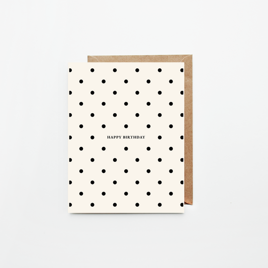 A  birthday card with a black polka dot pattern on an off-white background, featuring the text 'HAPPY BIRTHDAY' in the center. It is shown with a brown kraft envelope behind it on a clean white background.