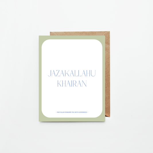 A greeting card with a white center panel, pale sage green border, and blue text reading 'JAZAKALLAHU KHAIRAN' with smaller text below, shown with a brown kraft envelope on a light gray background.