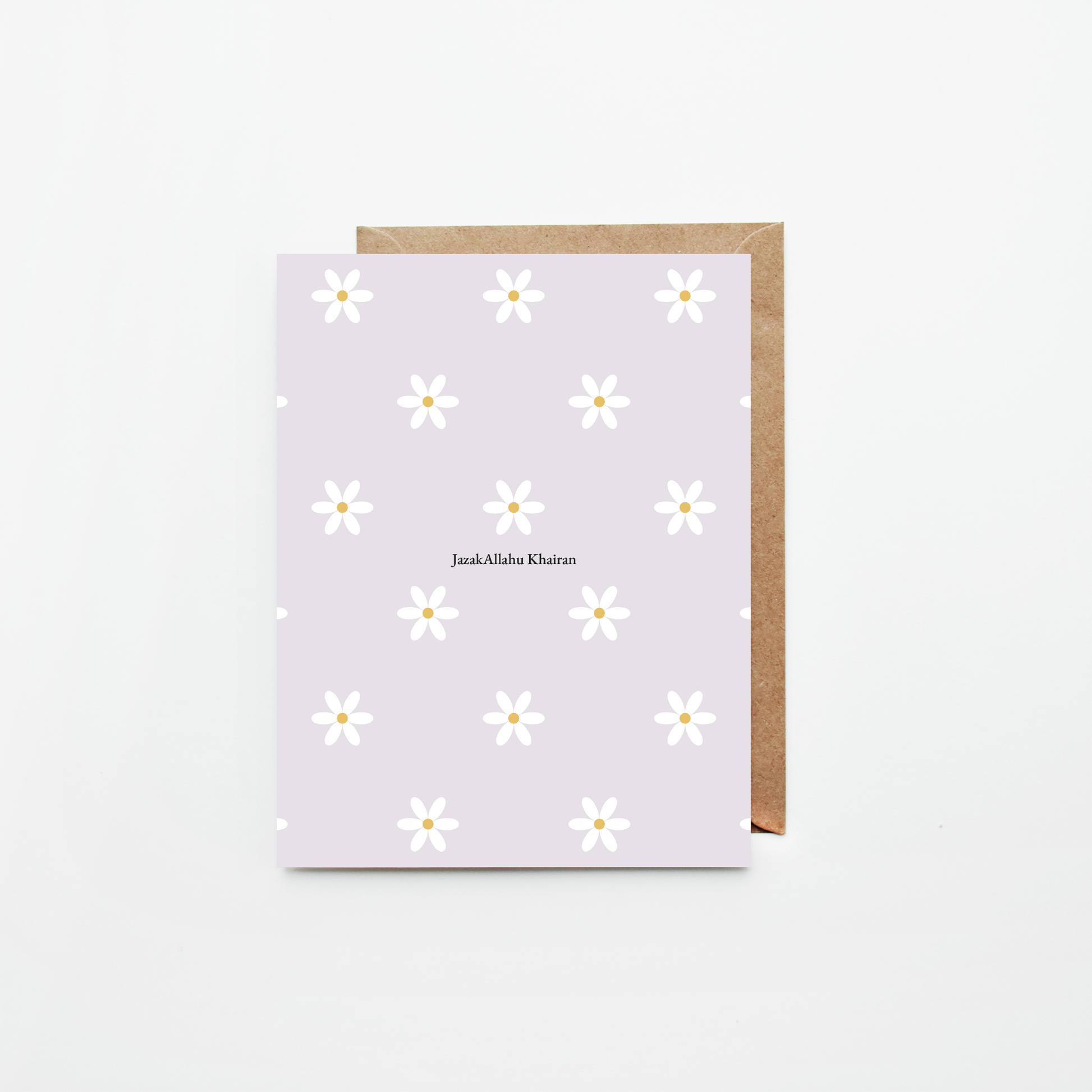 A greeting card with a light lavender background, white daisy pattern, and the name 'JazakAllahu Khairan' printed in the center, shown with a brown kraft envelope on a clean white background.
