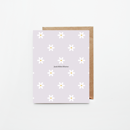A greeting card with a light lavender background, white daisy pattern, and the name 'JazakAllahu Khairan' printed in the center, shown with a brown kraft envelope on a clean white background.