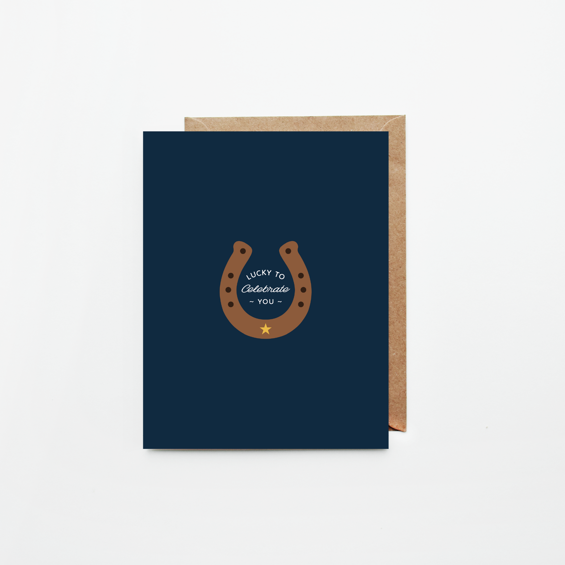 A navy blue greeting card with a brown horseshoe graphic and the words 'Lucky to Celebrate You' in white, shown with a brown kraft envelope on a light gray background.